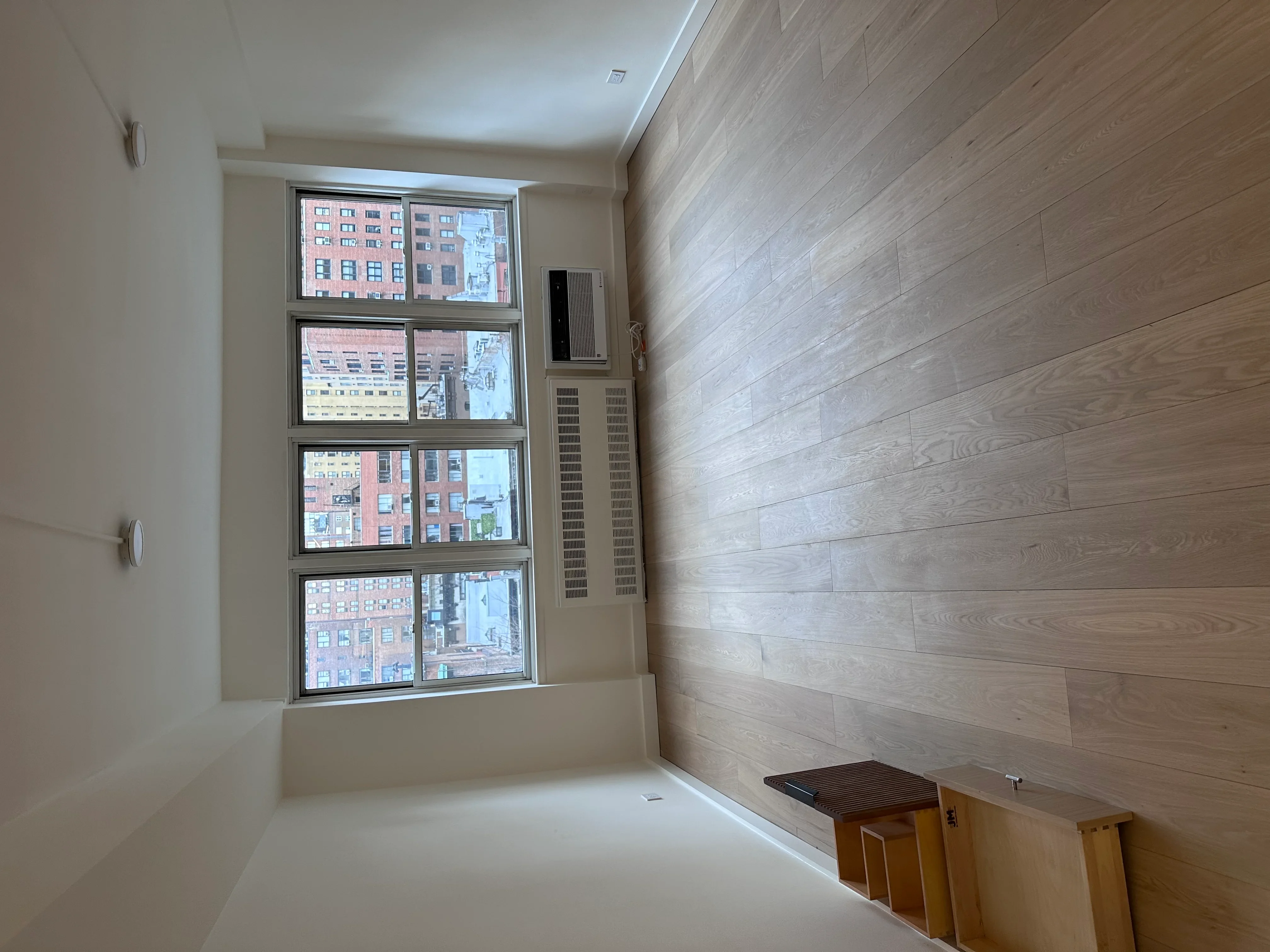Modern apartment interior with diagonal wood-look flooring and large windows overlooking city buildings